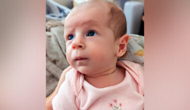 A baby in a pink onesie with floral embroidery gazes up and to the left while lying on a dinosaur-patterned blanket.