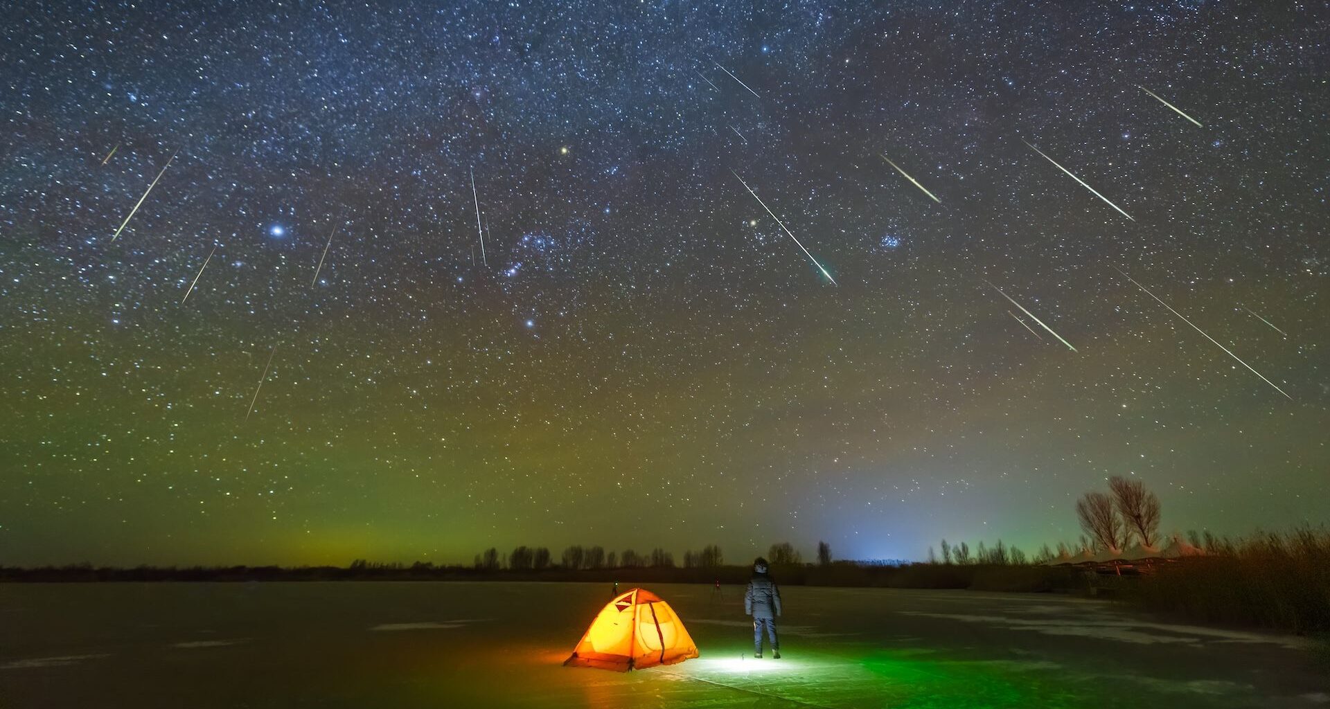 a camper stands outside a tent as the night sky is lit up by a meteor shower