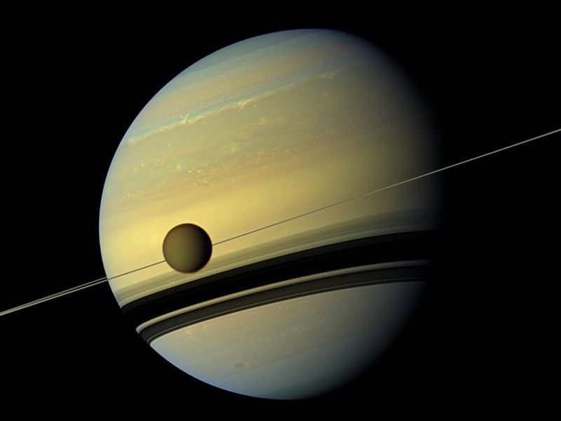 Yellowish orb of Saturn with a thin ring but shadows appearing thicker and the moon Titan.