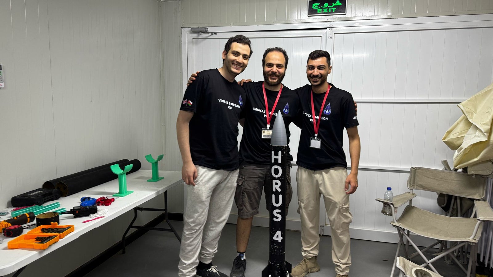 Three individuals, wearing matching black shirts with logos, stand together smiling in a room with a white interior. They are positioned next to a small model rocket labeled 'HORUS 4'. A table nearby displays various tools and rocket components, with folding chairs seen in the background.