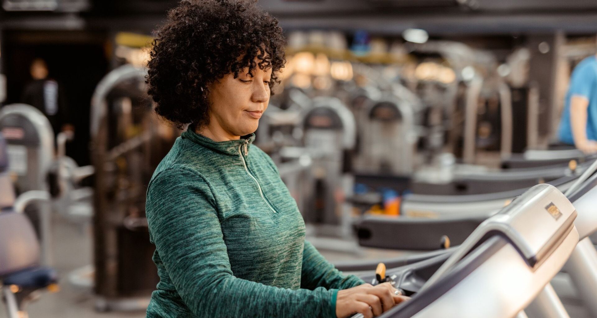 woman in a green long sleeved top on a treadmill and adjusting the settings; the camera shot is from the waist up with a gym setting behind her.