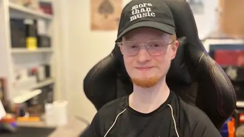 Max Corrigam is sitting in a leather chair inside a well lit bedroom. He is wearing a black hat which says "more than music" on it and is wearing glasses.