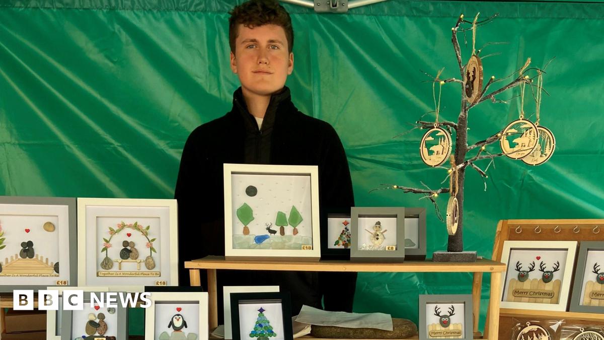 Aiden Thompson at is stall at Carlisle Teenage Market. His goods, including framed Christmas cards a pictures, are laid out on tables in front of him. He is wearing a black jumper and has dark hair and blue eyes.