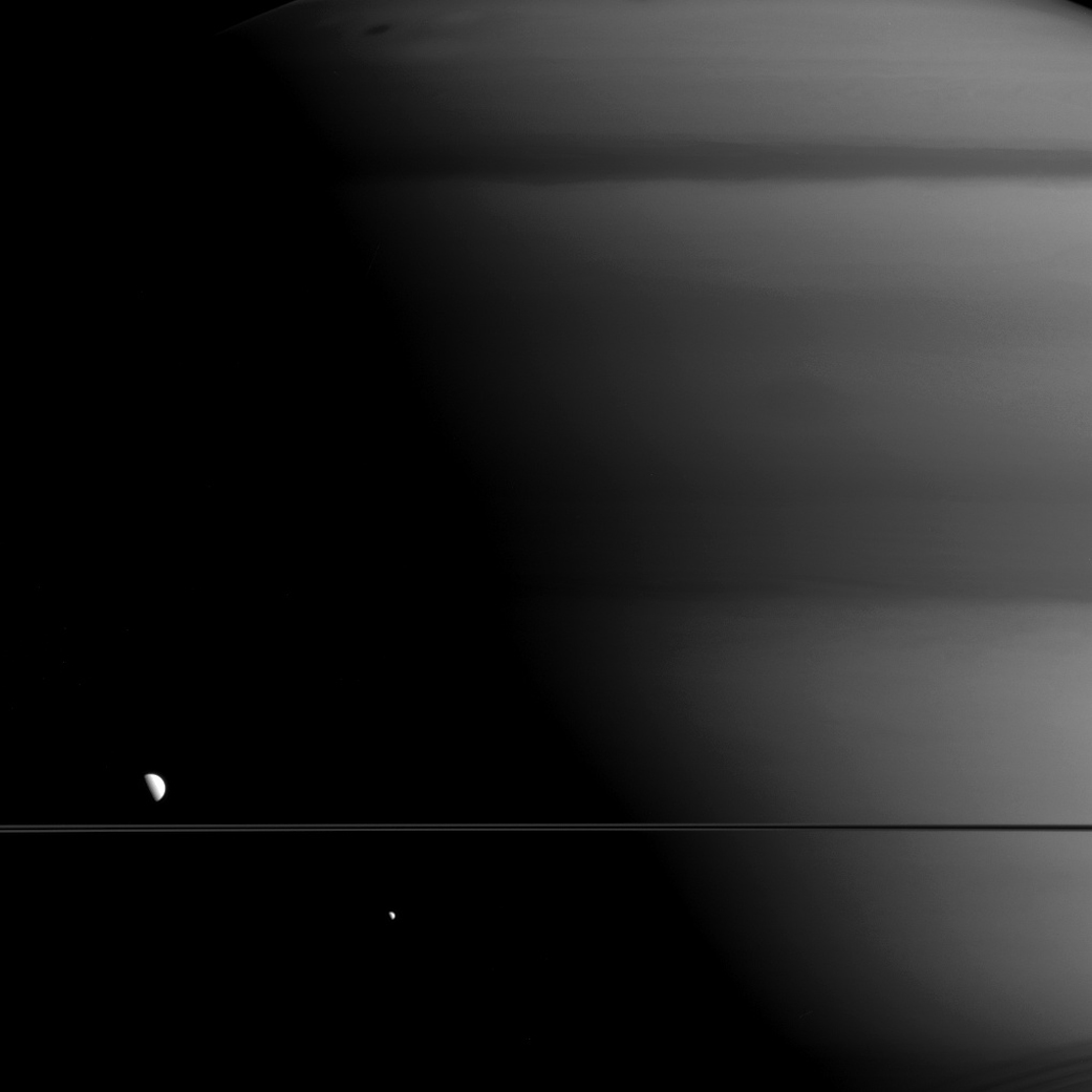 A black and white view of a huge planet with a line in the bottom right across the screen representing the planet's rings. There are two small white dots, a larger one above the ring to the left and a smaller one below the ring to the right.