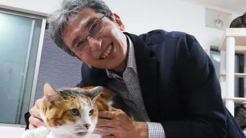 Hiroyuki Sasaki/Kyushu University Professor Sasaki smiles and leans down towards the table where he is holding a calico cat, mixed orange and brown fur with intently staring green eyes. They are in a cat rescue centre with other cats on a play tower in the background