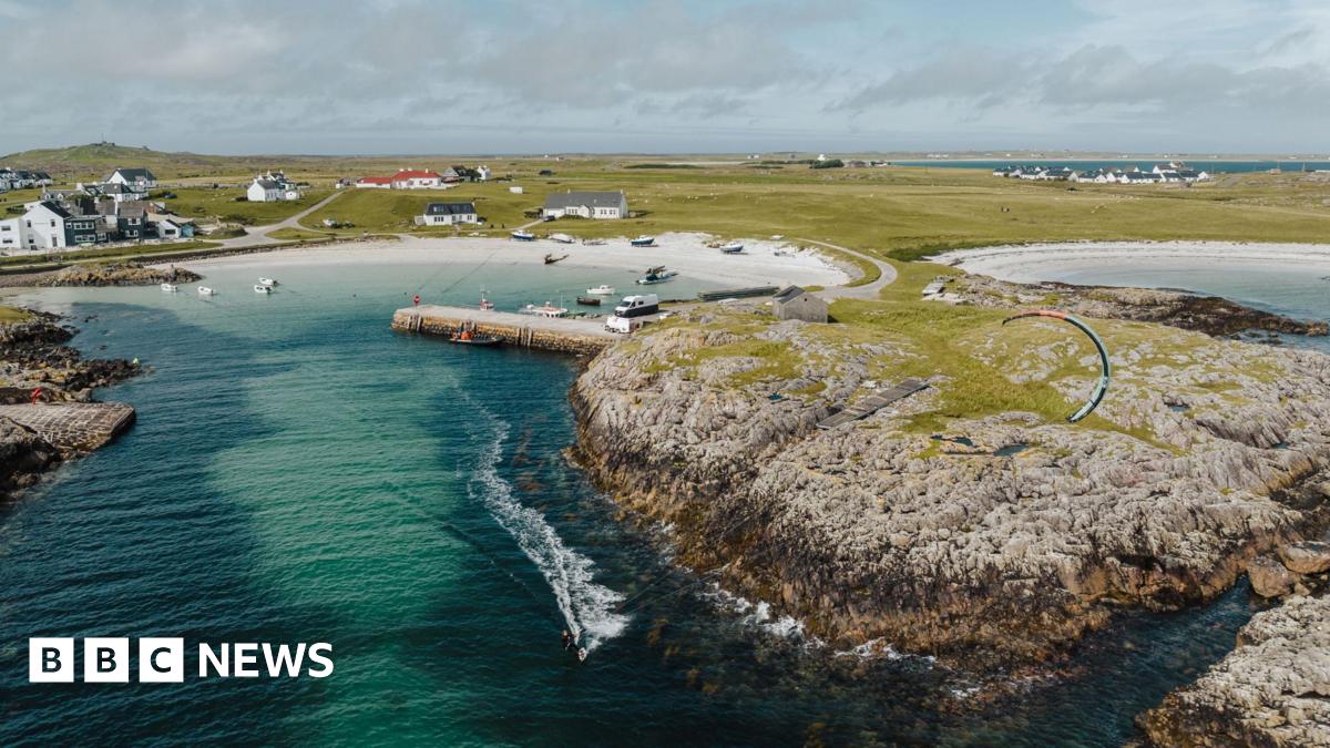 An aerial view of a community in Tiree. There are white-walled houses, a rocky shoreline and two beaches of white sand. The sea is turquoise and a dark green. A kite surfer leaves a frothy wake on the sea's surface as they travel out from a small bay.