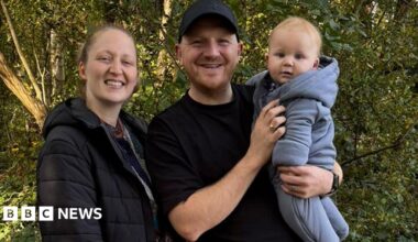 A man and woman are standing together in an outdoor setting. The woman on the left is wearing a dark jacket and is smiling a the camera. The man is wearing a black shirt and cap is also smiling and is holding a baby. The baby is dressed in a light-coluored hooded baby grow. Behind them are trees and foliage.