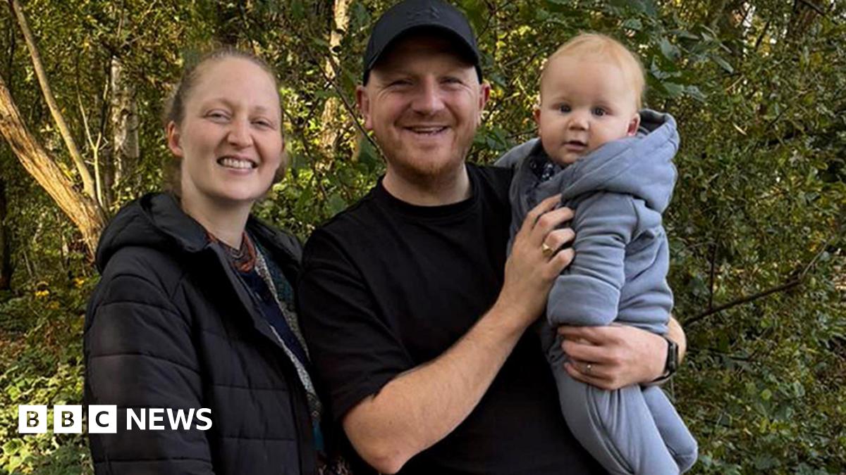 A man and woman are standing together in an outdoor setting. The woman on the left is wearing a dark jacket and is smiling a the camera. The man is wearing a black shirt and cap is also smiling and is holding a baby. The baby is dressed in a light-coluored hooded baby grow. Behind them are trees and foliage.