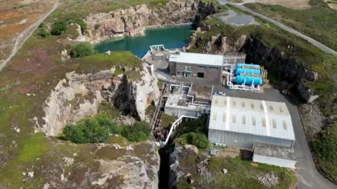 BBC A view from above shows Jersey's desalination plant, surrounded by cliff faces and made up of water containers, and two main buildings with an area of seawater to the top of the image. 