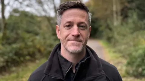 BBC A man standing on a path lined by trees and bushes. He has grey hair and a grey beard. He is wearing a black fleece.