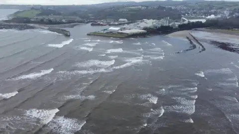Par Harbour is seen with waves heading toward the shore on which there are houses and buildings 