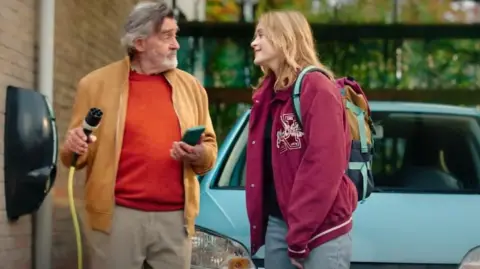 Zet ook de knop om A man with an electric-charging cable stands in front of an electric car with a young woman