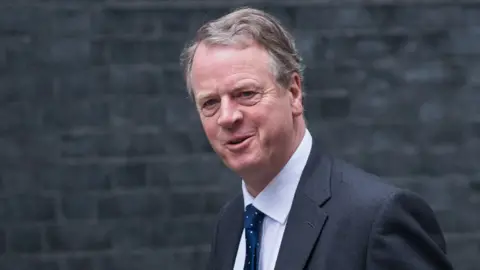 Getty Images Alister Jack, who has dark grey hair, looks at the camera while walking in front of a grey brick wall. He is wearing a dark grey suit, white shirt and blue tie. 