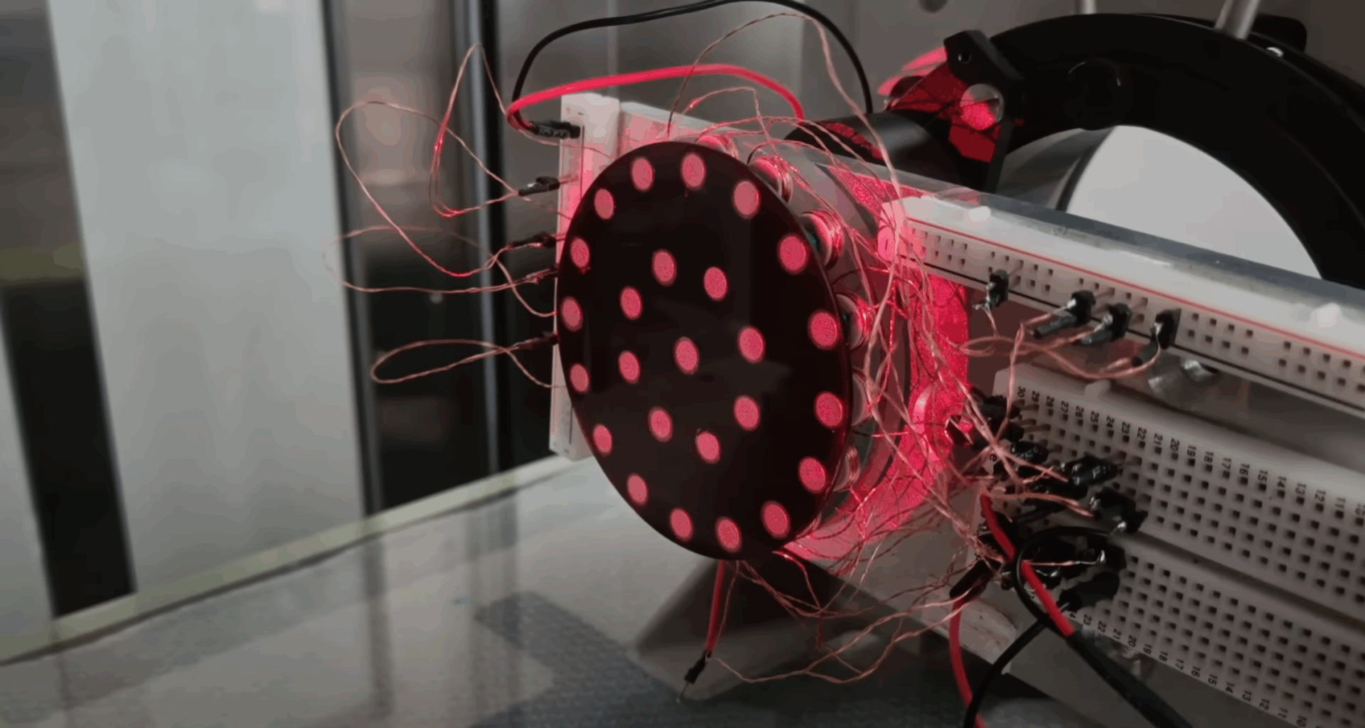 A vertically-mounted black disk with a concentric pattern of reflective disks is illuminated under a red light. A large number of copper wires run away from the the disk to a breadboard.