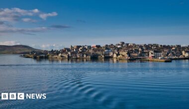 Houses and other buildings on the waterfront at Lerwick. There are ripples on the surface of the sea and the sky is blue.