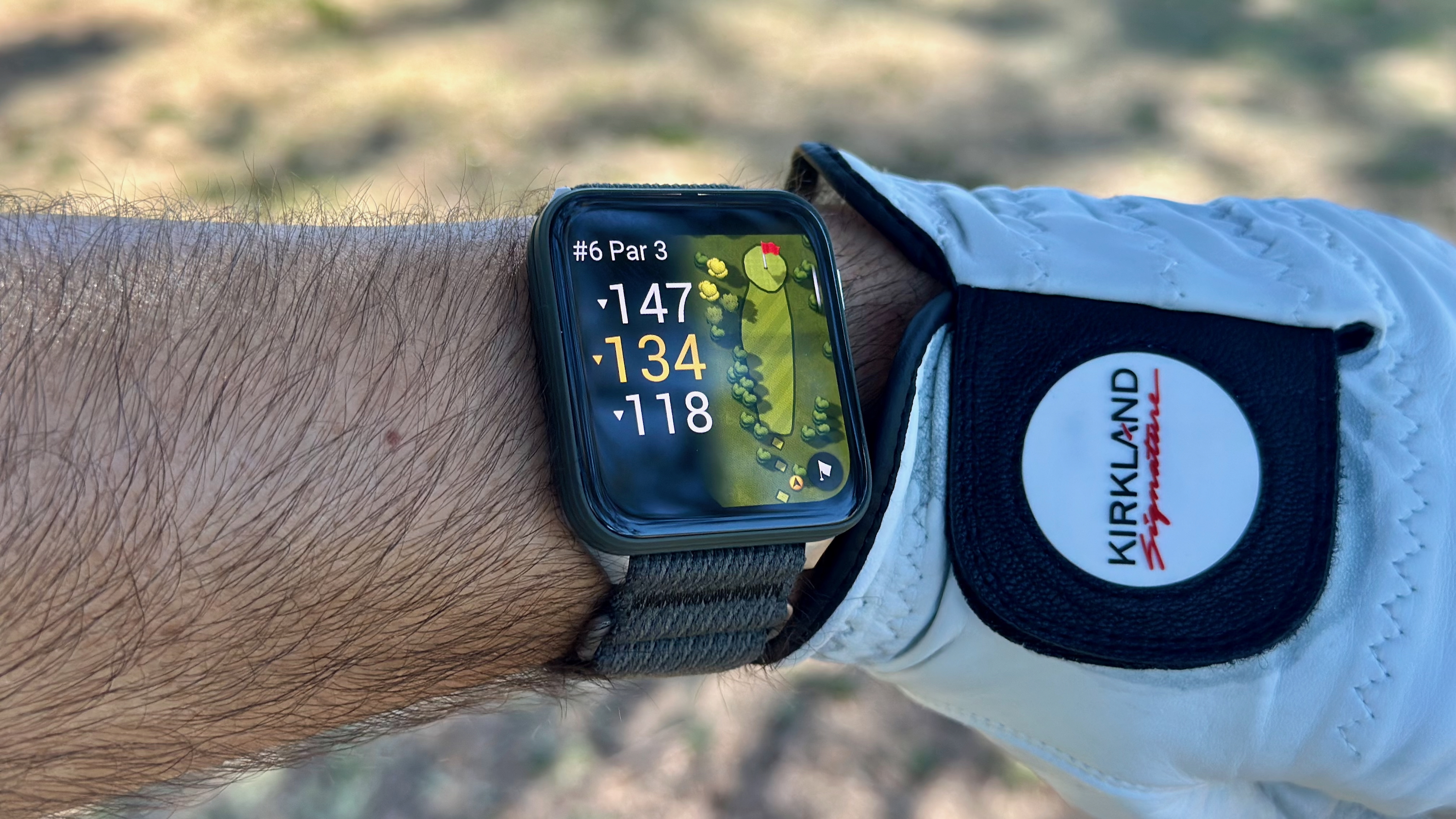 The Garmin Venu X1 on a wrist next to a golf glove, showing the yardage for the hole with fairway rough in the background.