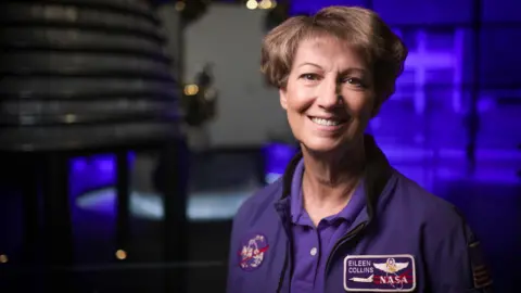 Tony Jolliffe/ BBC News Eileen Collins wearing a blue jacket with a round Nasa patch on one side and a rectangular shuttle patch on the other with her name. She has short brown hair and brown eyes and is smiling as she looks directly to camera. She is in the space gallery at the Science Museum and an out of focus rocket engine can be seen behind her. 