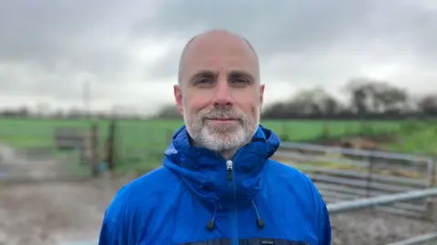 Laurence Couldrick is in a blue waterproof stands in front of a farm gate. There are green fields in the background. 