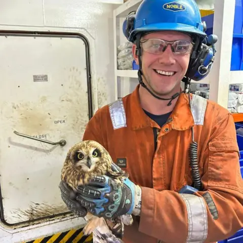 Sam Crowe Offshore worker Sam Crowe in orange overalls, smiling at camera, and holding an owl.