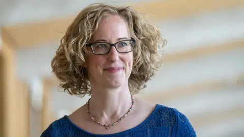 PA Media Lorna Slater, who has blonde curly hair and glasses, smiles as she looks to her left. She is wearing a blue top and multi-coloured necklace.