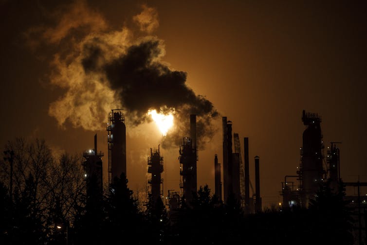 a nighttime image of tall flare stacks at a refinery. a bright flame emerges from one along with a large plume of smoke