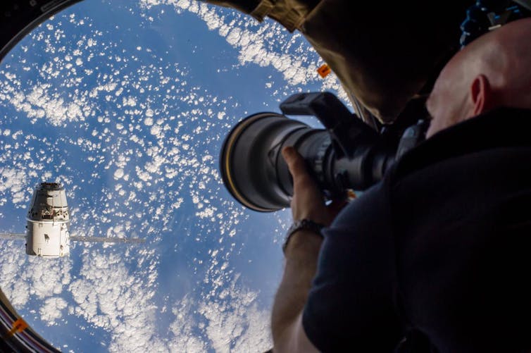 An astronaut pointing a camera out a circular window in the International Space Station at a