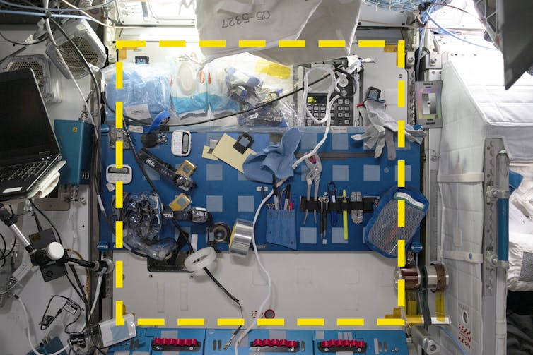 The wall of a space station module. In the center is a blue metal panel with 40 pieces of Velcro arranged in a grid. More Velcro is visible on the wall. Many different items are stuck to the wall. A yellow square is superimposed on the central part of the image.