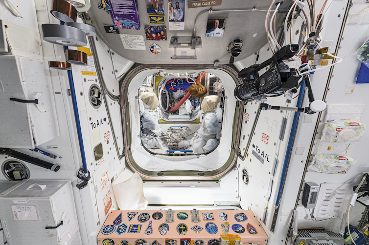 A view from one module of a space station into the airlock. In the airlock are two spacesuits facing each other. At the threshold, there is a hatch door at the top with pictures of people and other items on it. Below is a salmon-colored bulkhead with stickers of mission patches on it.
