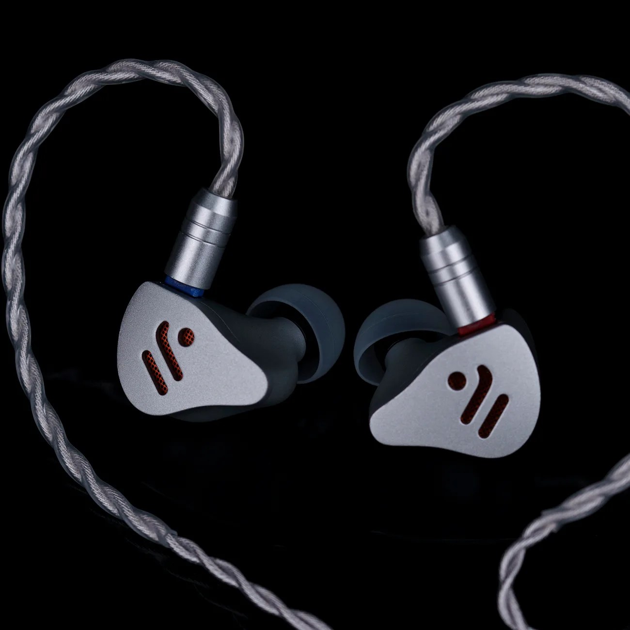 A pair of in-ear monitors with silver faceplates featuring a red logo and black ear tips. They are connected by a braided silver cable with metal connectors, one marked with a blue ring and the other with a red ring, against a black background.