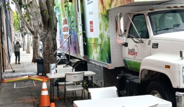 A mobile health clinic truck is parked on a city street beside folding tables and chairs, with orange traffic cones placed nearby.