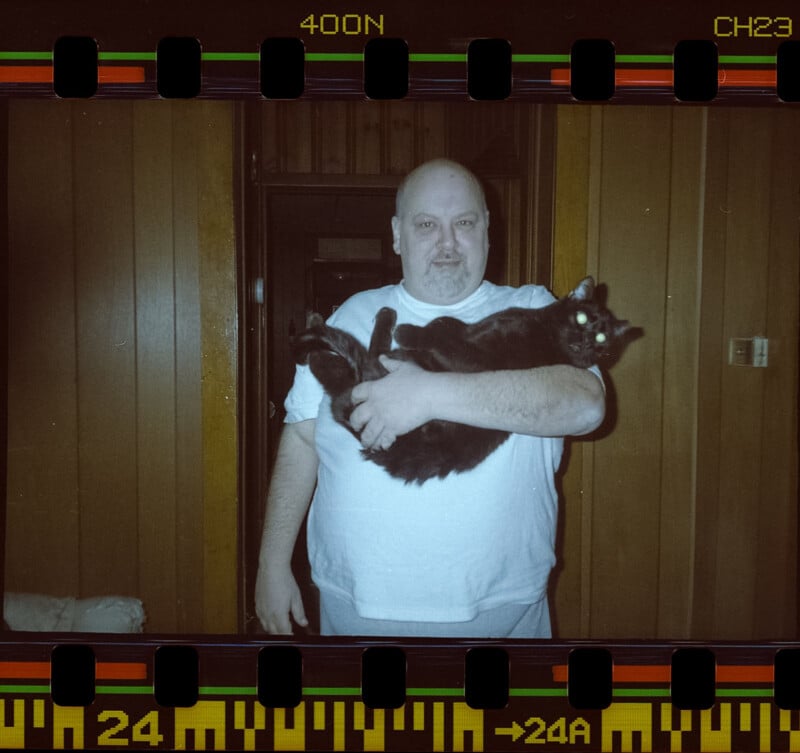 A man in a white t-shirt stands indoors, holding a large black cat with glowing eyes. The setting has wood-paneled walls, and the image has a filmstrip border effect.