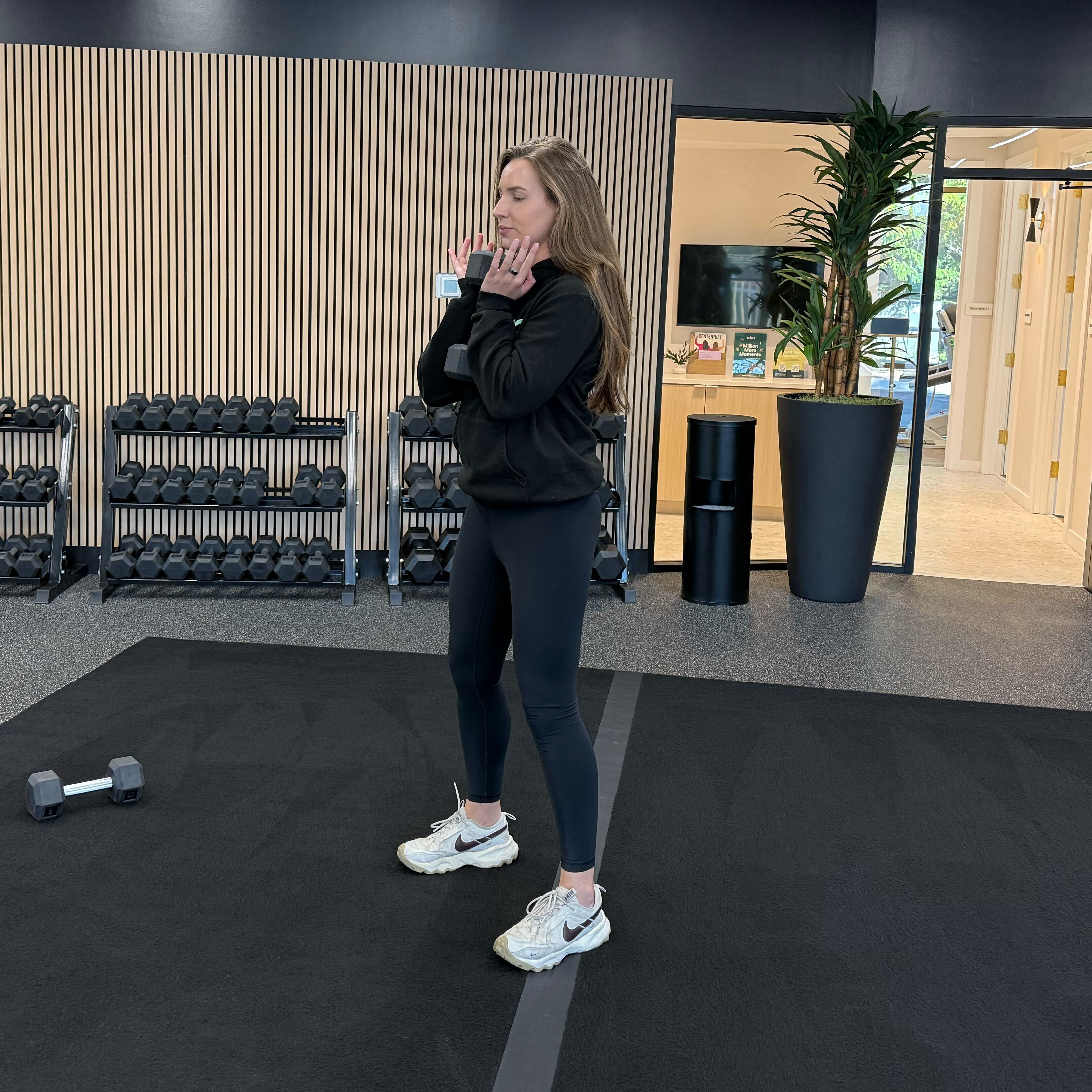 Felicia Hernandez, a personal trainer for Eden Health Club, stands holding a dumbbell beneath her chin in a fitness studio.