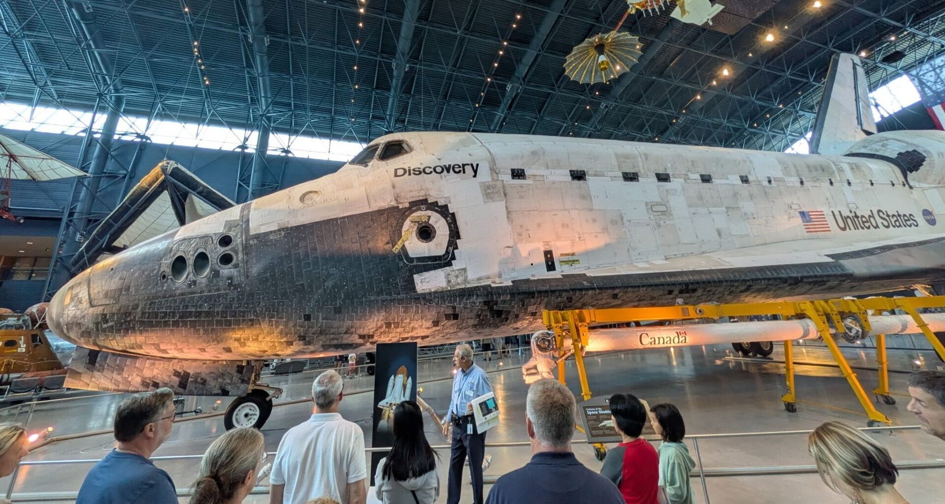The space shuttle Discovery, a white and black shuttle is on display at the Smithsonian Museum where people walk around it and view it.