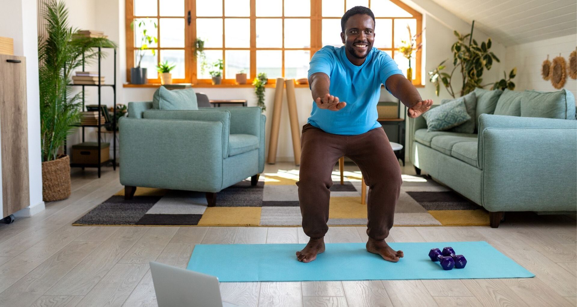man performing a squat with arms outstretched in front on a blue exercise mat. there's a laptop on the floor in front of him and a living room setting with two sofas and a large window behind him