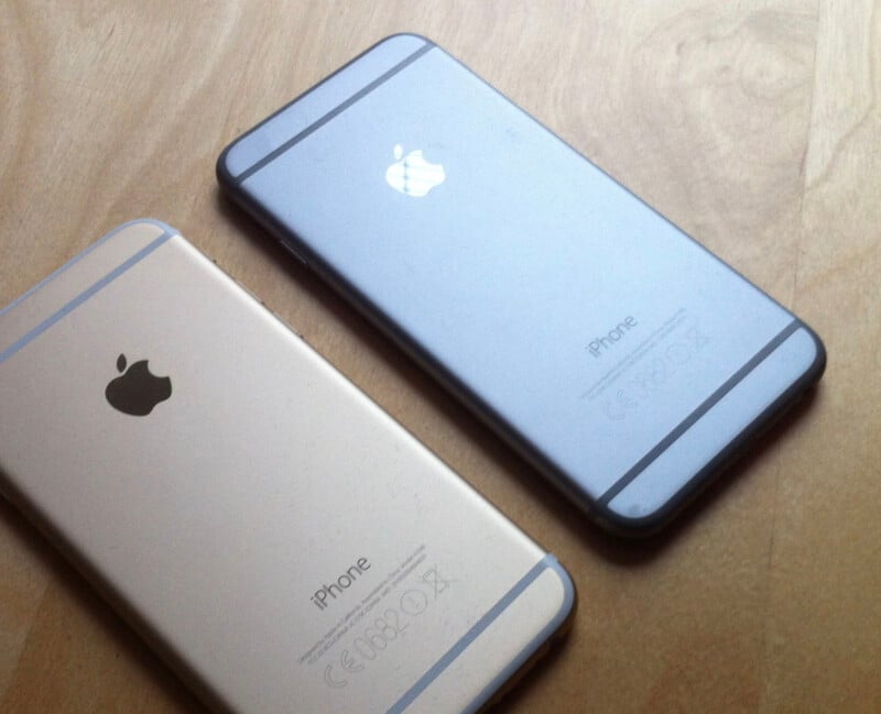 Two iPhones are lying face down on a wooden surface. One is gold and the other is gray, both showing the Apple logo and the word "iPhone" on their backs.