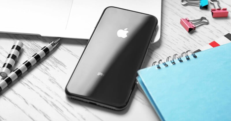 A black iPhone lying face down on a white wooden desk next to a silver laptop, a striped pen, a blue spiral notebook, and colorful binder clips.