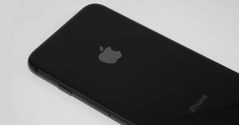 A close-up of a black iPhone placed on a white surface, showing the Apple logo and part of the word "iPhone" on the back.
