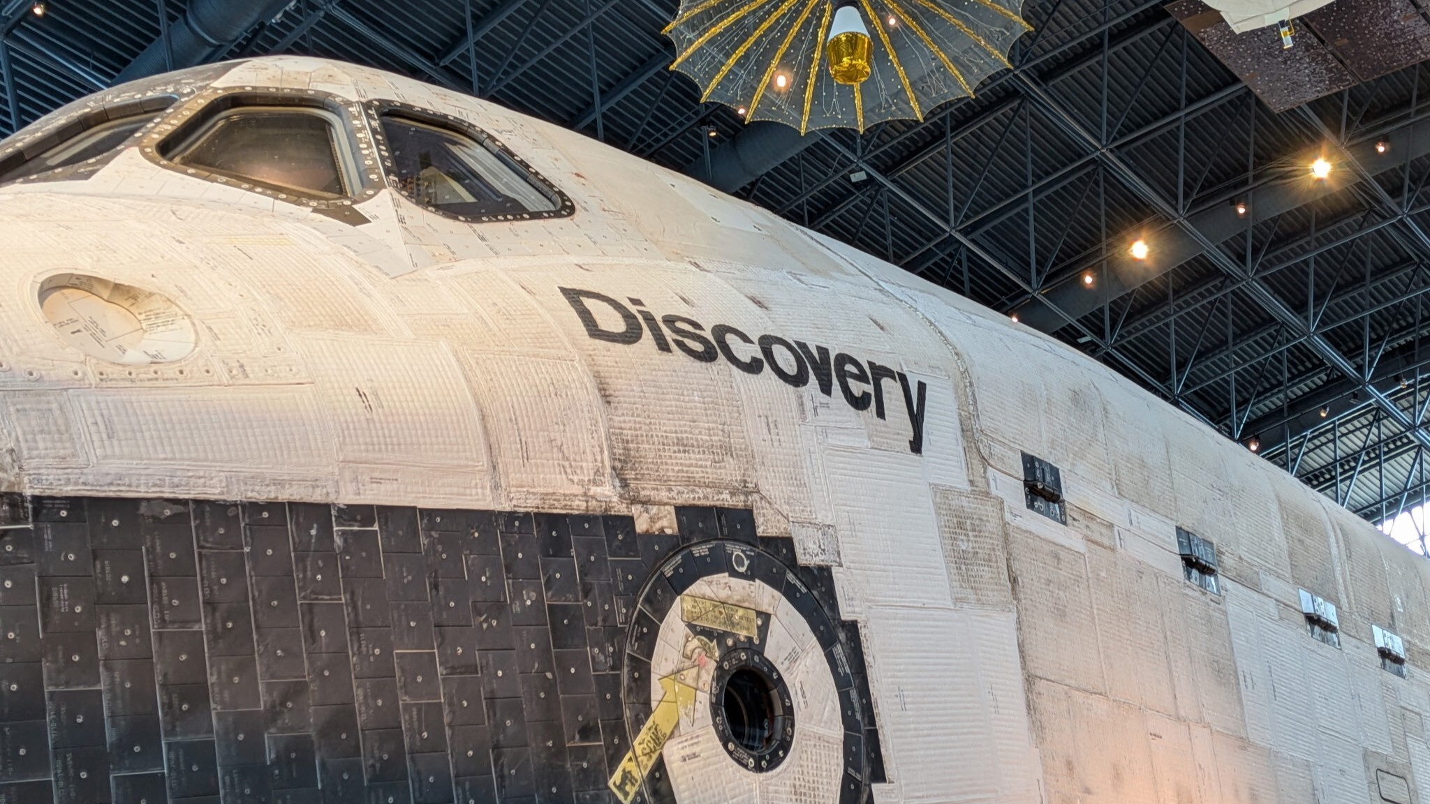 The space shuttle Discovery, a white and black shuttle is on display at the Smithsonian Museum where people walk around it and view it.