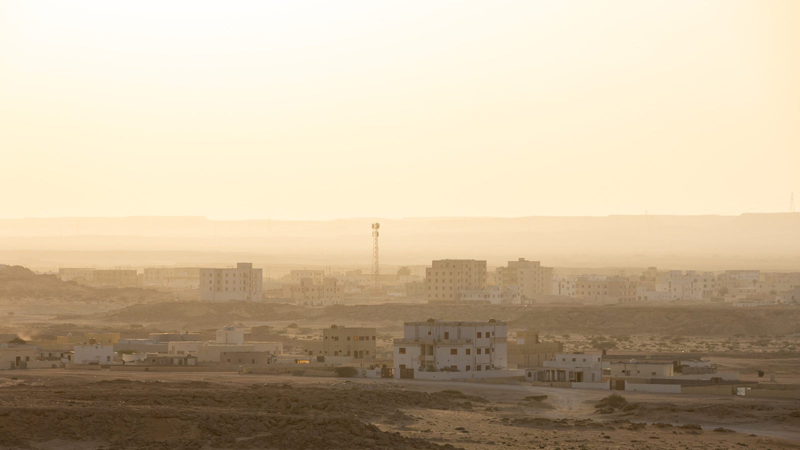 A hazy desert landscape at sunset, featuring low-rise buildings in the foreground and a distant horizon, with a cell tower visible among the structures.