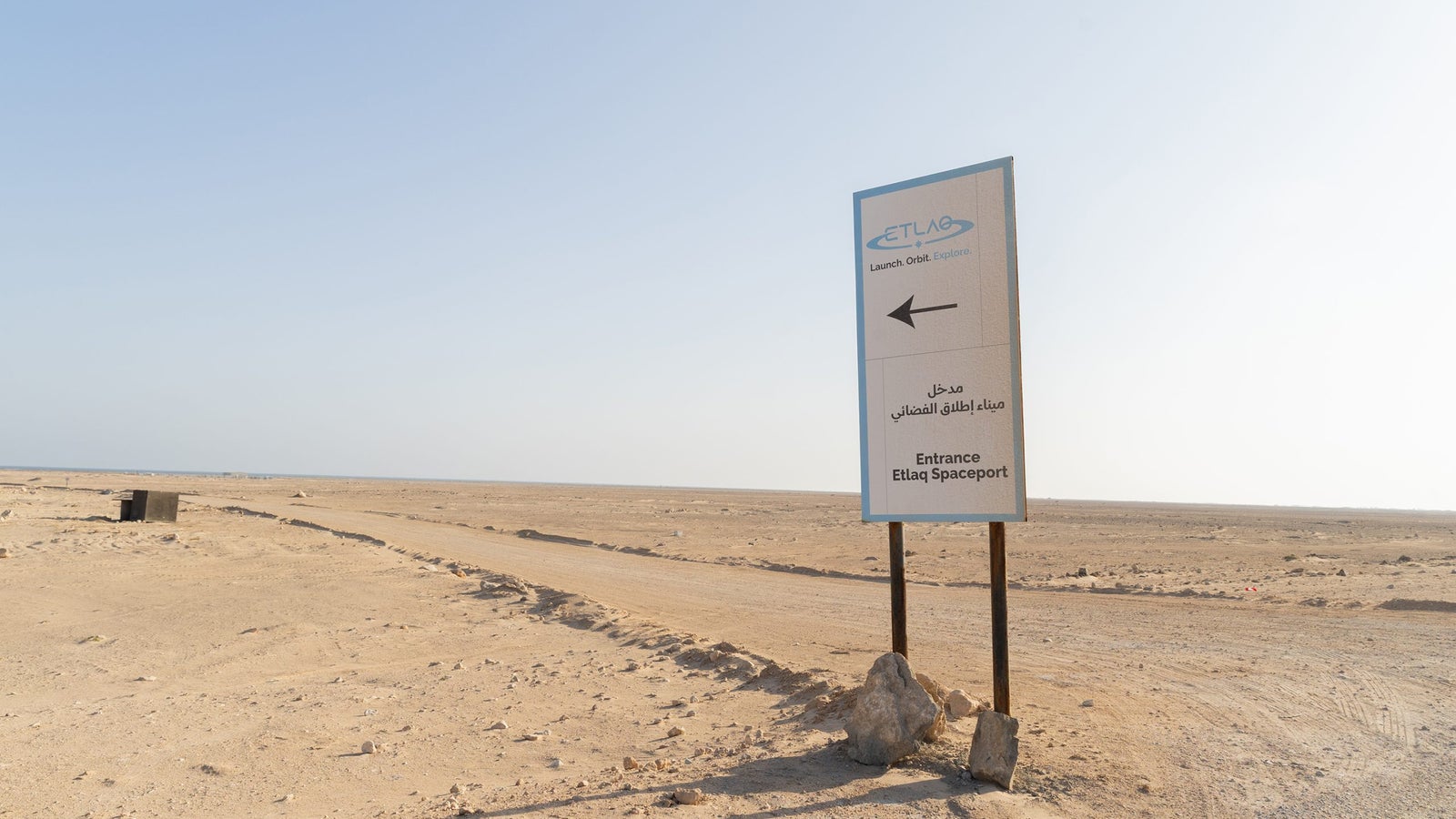 A sign directing towards the entrance of Etlaq Spaceport, standing on a sandy desert landscape with a clear sky in the background.