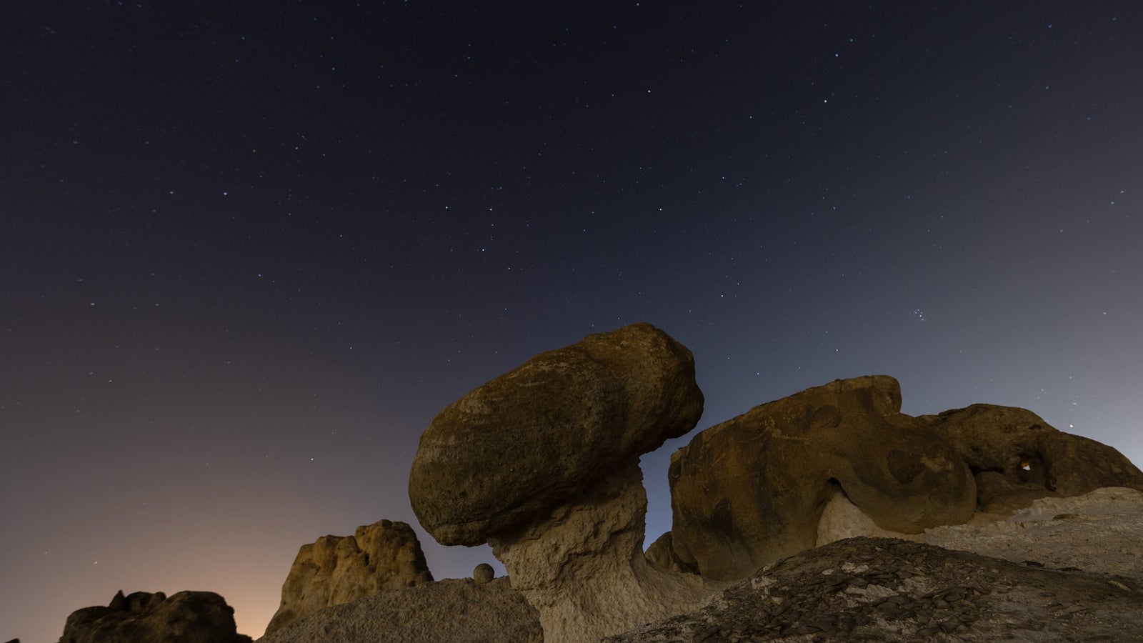 A nighttime landscape featuring large rock formations under a starry sky, with a prominent mushroom-shaped rock in the foreground and hints of light from a distant horizon.