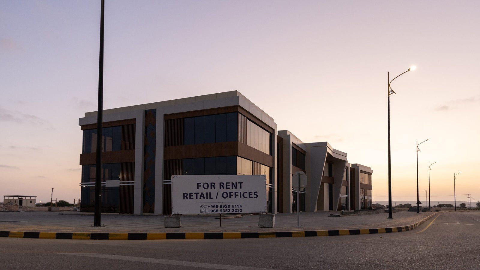 A modern two-story commercial building with large glass windows and decorative elements, featuring a prominent sign stating 'FOR RENT RETAIL / OFFICES.' The image is taken during twilight, with streetlights illuminating the area and a clear sky in the background.