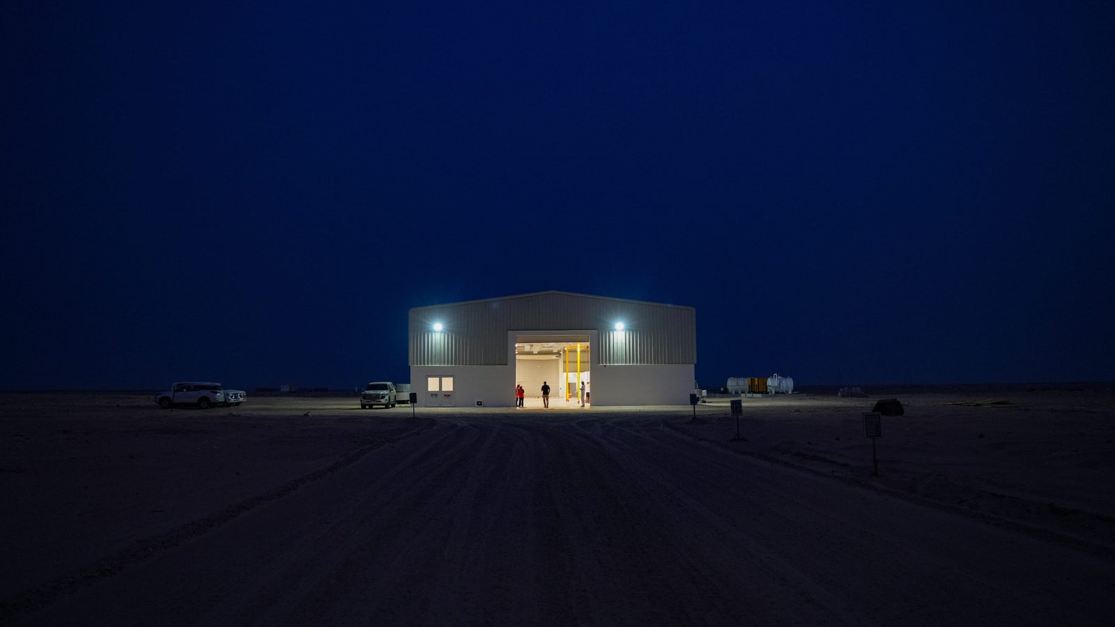 A dimly lit building in a barren landscape during dusk, with open doors revealing light inside and a few figures standing outside; two vehicles parked nearby and a clear sky transitioning from blue to black.
