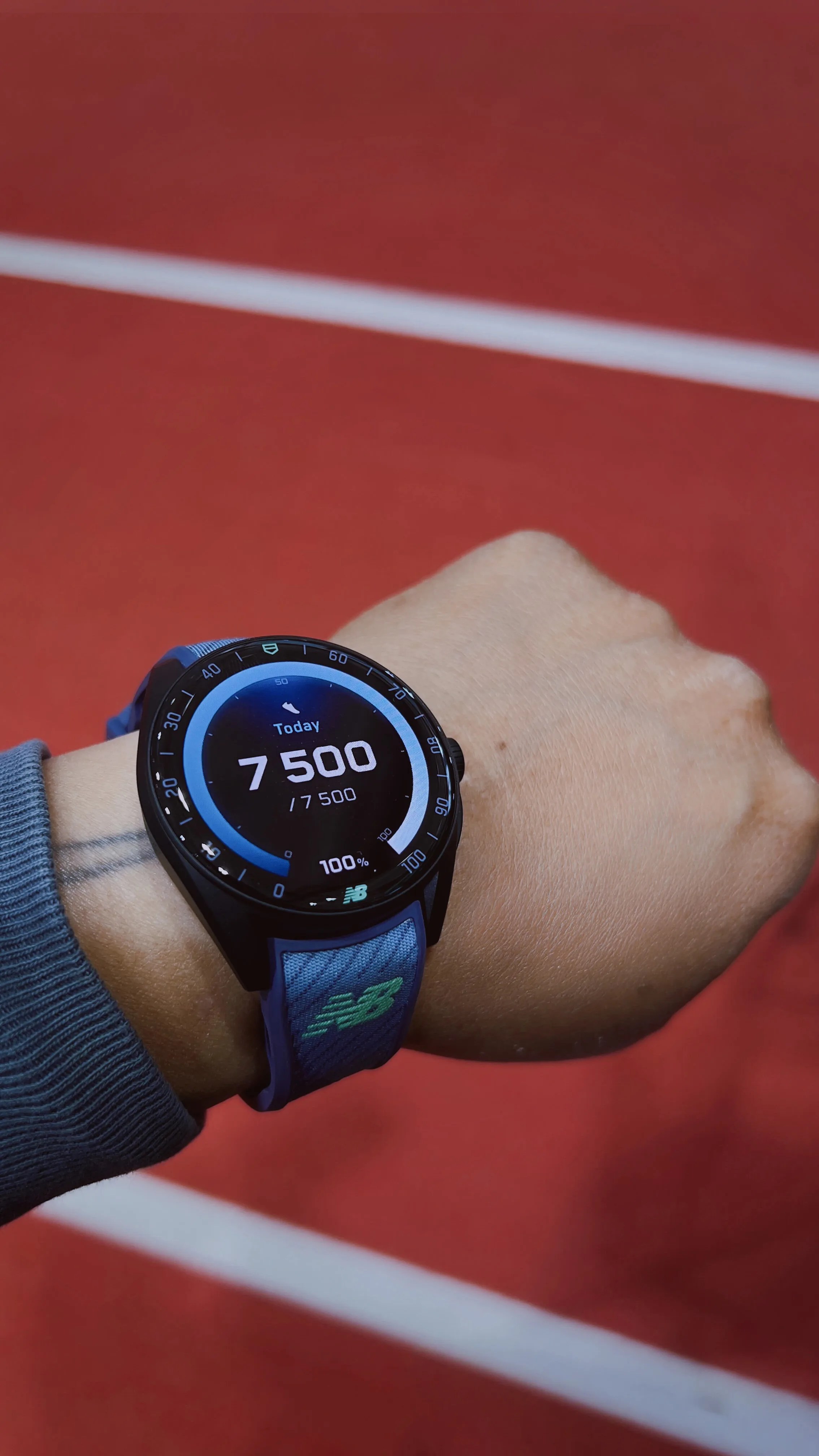 Smartwatch with a blue New Balance strap showing 7,500 steps on a wrist against a red track background.