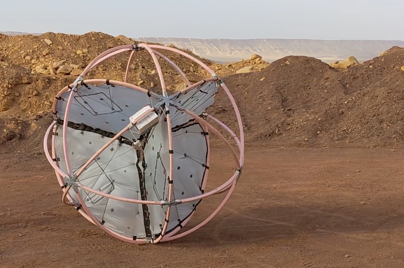 Tumbleweed rovers: spherical object composed of interlocking loops, with rounded flat panels inside it. It is sitting near mounds of dirt in a desert.