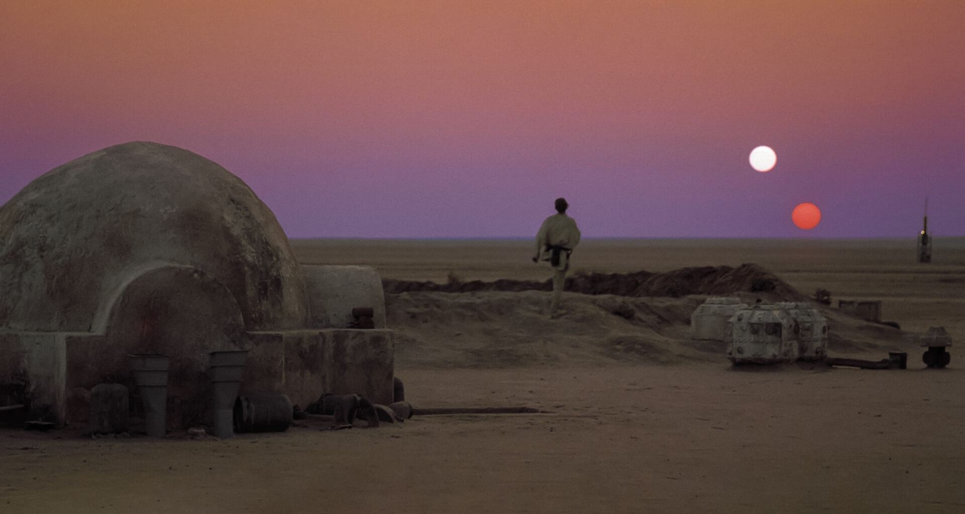 A man wearing white stands outside a stone igloo in the middle of a desert with a red and white sun in the dusky red sky.