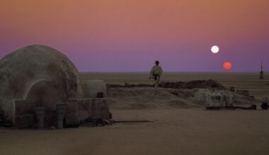 A man wearing white stands outside a stone igloo in the middle of a desert with a red and white sun in the dusky red sky.