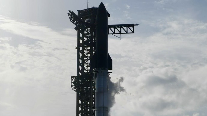 A black tiled upper stage and chrome lower stage rocket stands against a cloudy sky and dark launch tower.
