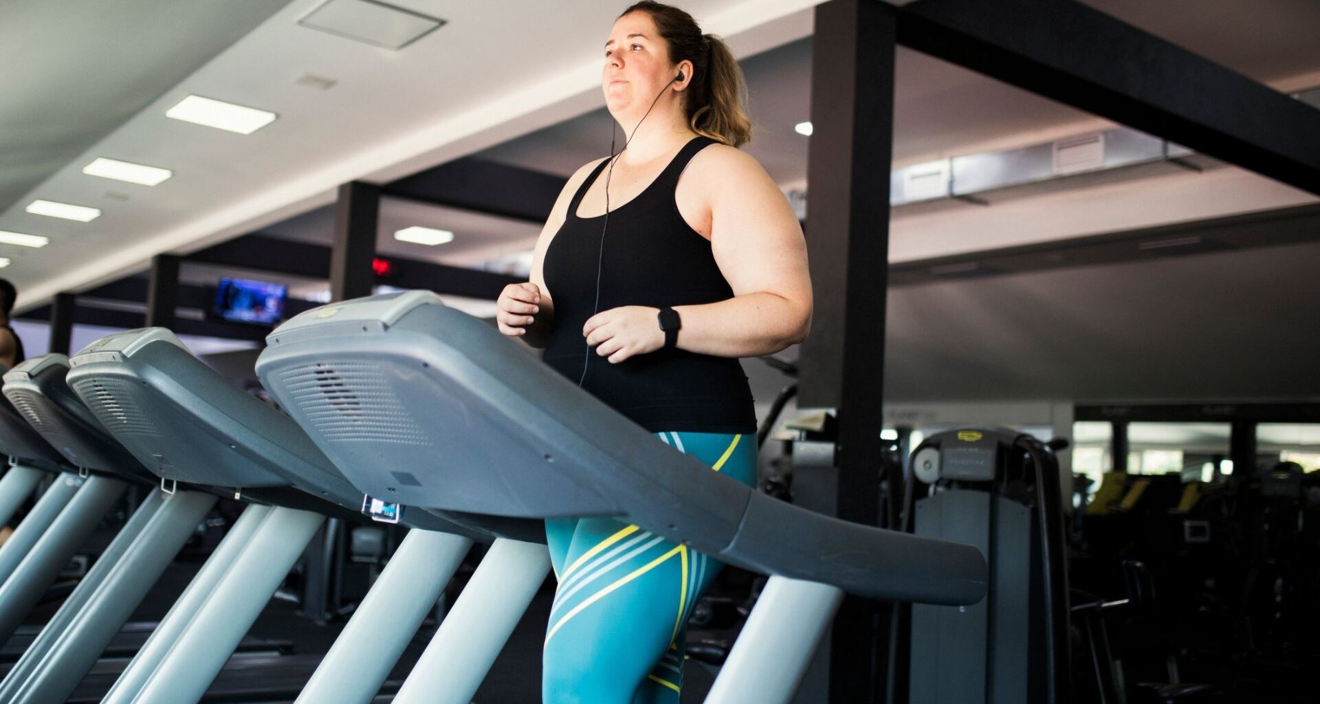 woman walking on a treadmill with earphones in, she's in a gym setting wearing turquoise leggings and black vest.