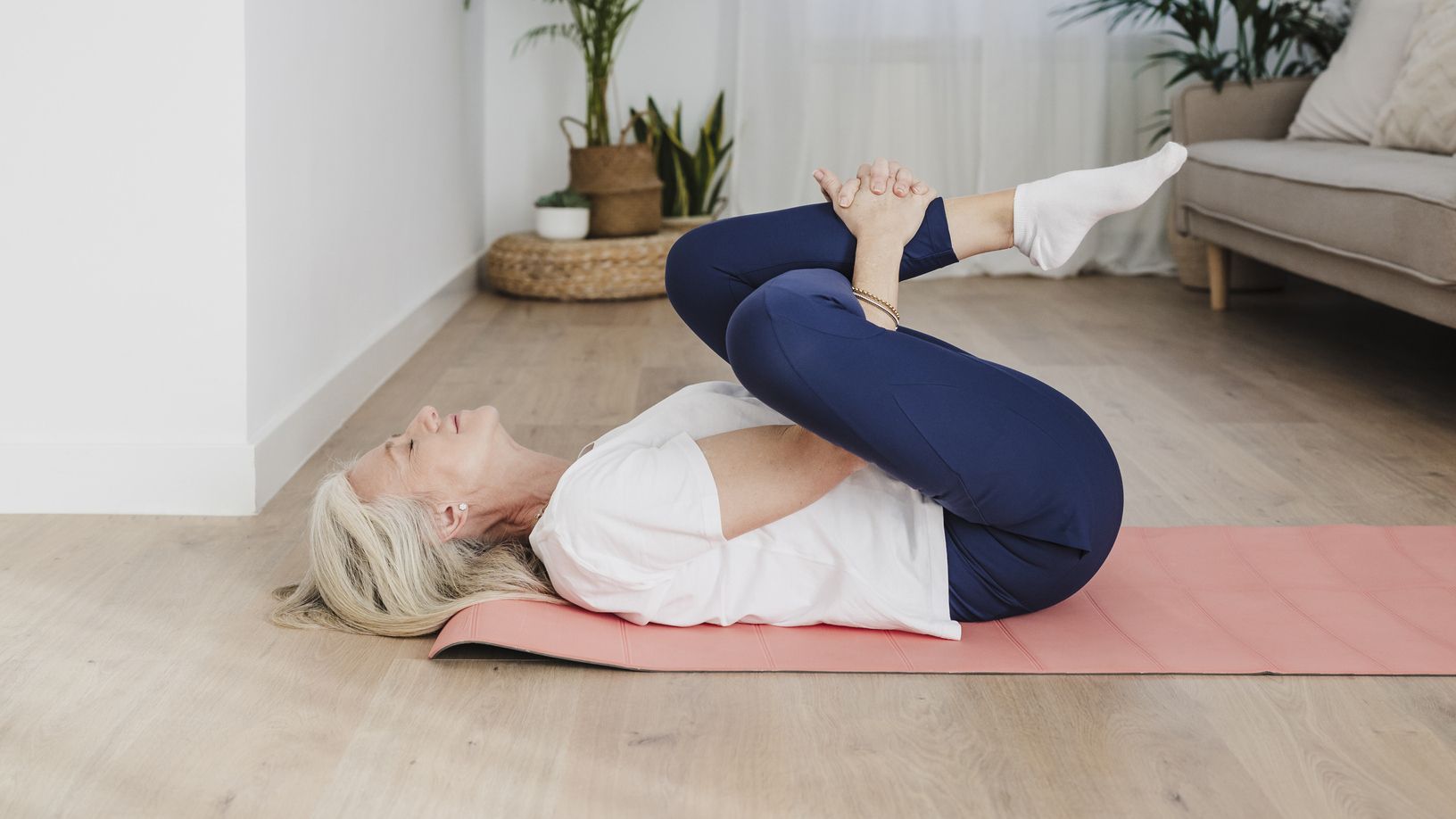 Woman lies on a yoga mat on her back in a living room, performing a leg stretch